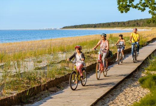 Eine vierköpfige Familie fährt mit dem Fahrrad auf dem Radweg am Strand entlang.