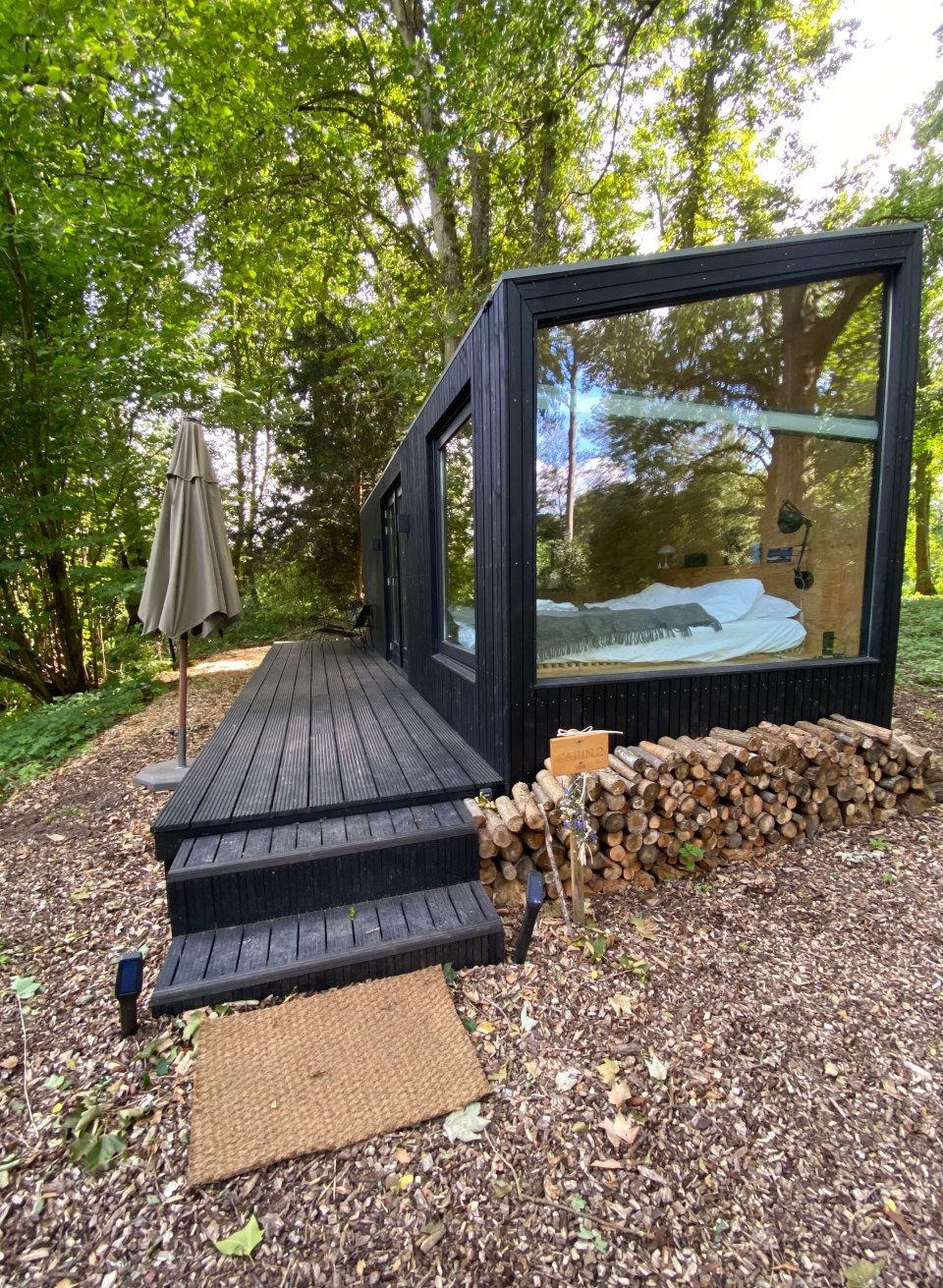 Schwarzes Tiny House mit Glasfront inmitten eines Waldes bei Schloss Neuhoff, davor Holzstapel und Terrasse.