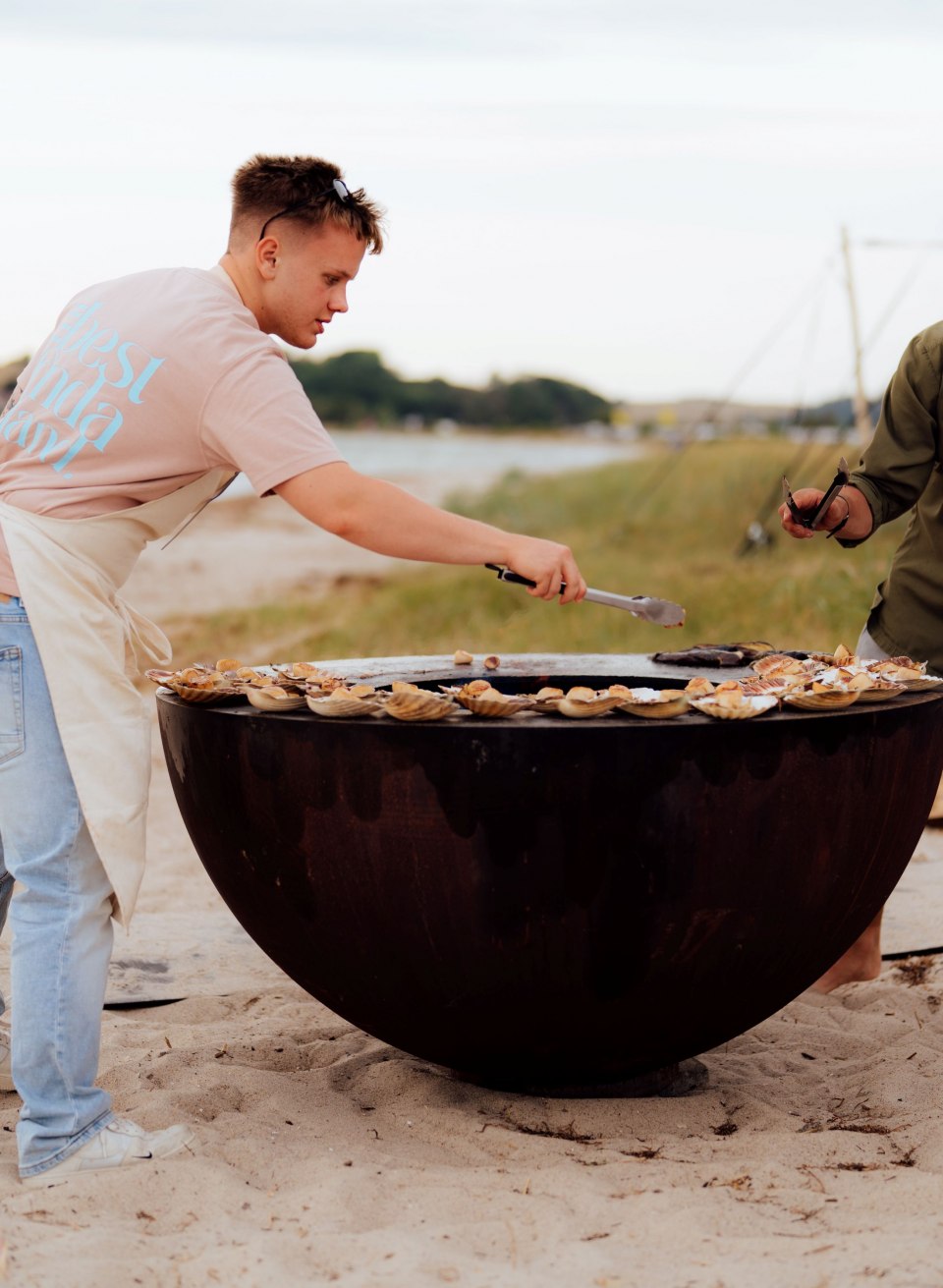 Zwei Männer grillen regionale Speisen auf einer großen Feuerplatte am Strand von Thiessow, dahinter Meer und Zelt. // Am Strand von Thiessow brutzeln frische regionale Speisen auf der Feuerplatte – Teamwork, Rauch und salzige Luft. // © MV-T/Petermann Zwei Männer grillen regionale Speisen auf einer großen Feuerplatte am Strand von Thiessow, dahinter Meer und Zelt.