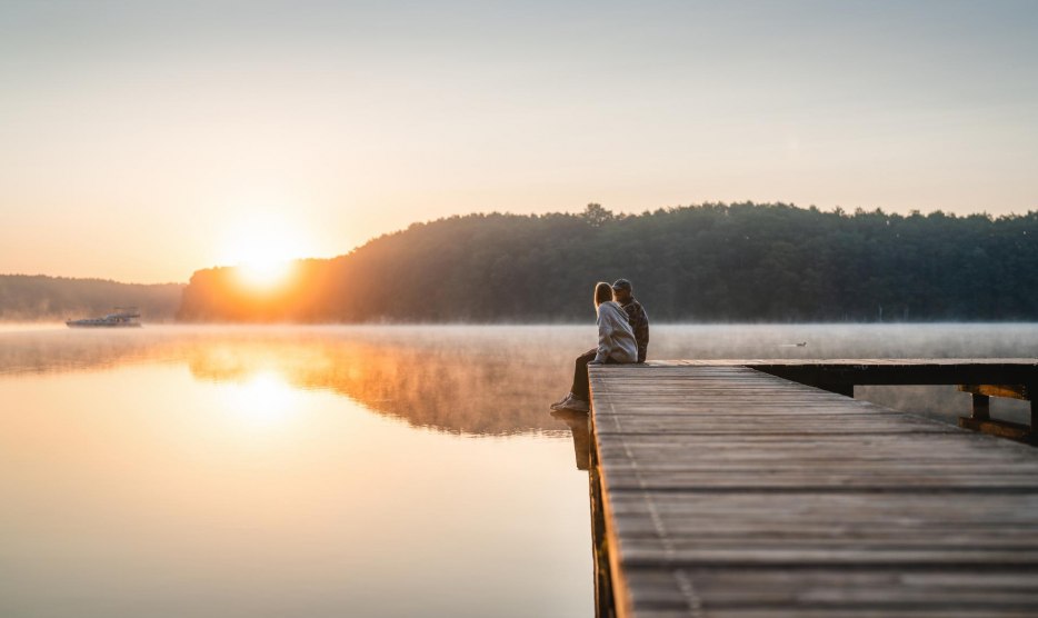 Sitzen auf dem Steg den Abend genießen am Mirower See, © TMV/Gross Ein Paar sitzt auf einem Steg zum Sonnenuntergang am Mirower See. Im Hintergrund fährt ein Hausboot vorbei.