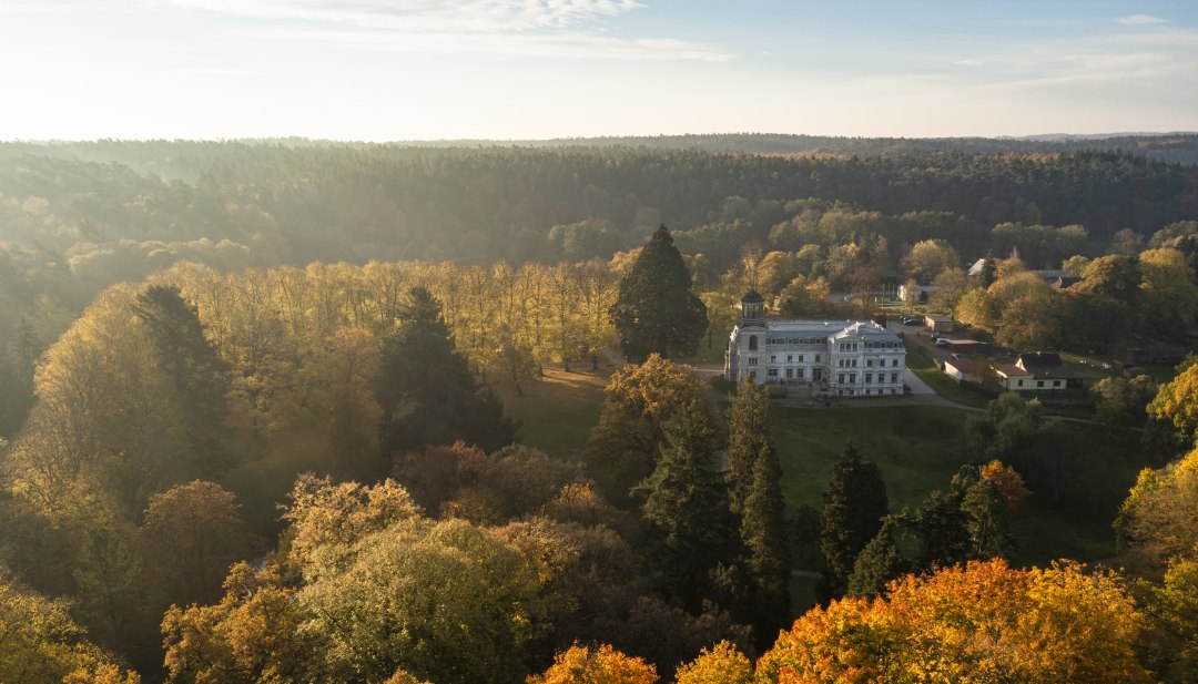 Herbstzauber im Schlosspark – das elegante Herrenhaus liegt eingebettet in bunte Baumkronen und lädt zum Spaziergang in traumhafter Naturkulisse ein., © Stefan von Stengel Luftaufnahme eines Schlosses inmitten herbstlich gefärbter Bäume, umgeben von einem weitläufigen Park und waldreicher Landschaft.