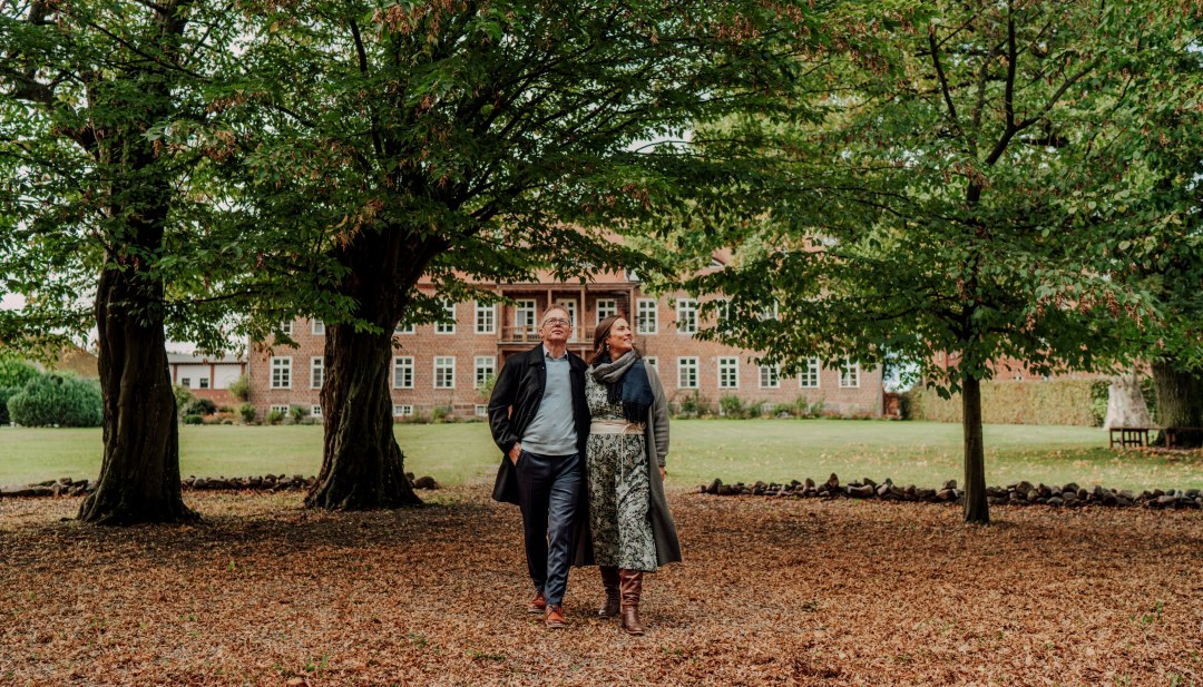 Een koppel wandelt onder grote bomen in het park, met het Romantik Hotel Gutshaus Ludorf op de achtergrond.