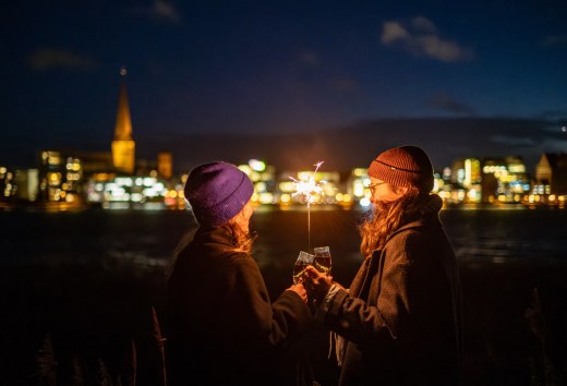 Zwei Personen sto&szlig;en mit Sekt an, w&auml;hrend sie eine Wunderkerze halten, mit der beleuchteten Skyline von Rostock in Mecklenburg-Vorpommern im Hintergrund.