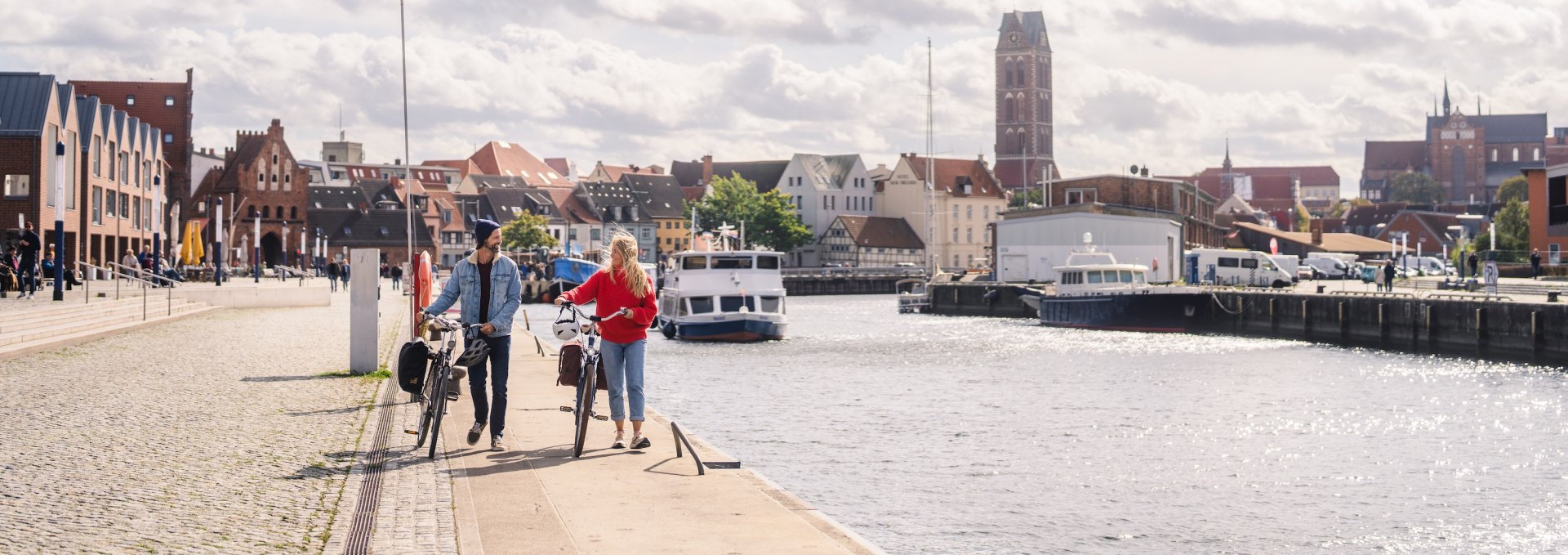 Ein Paar schiebt Fahrr&auml;der am alten Hafen von Wismar entlang, im Hintergrund die historische Skyline der Stadt.