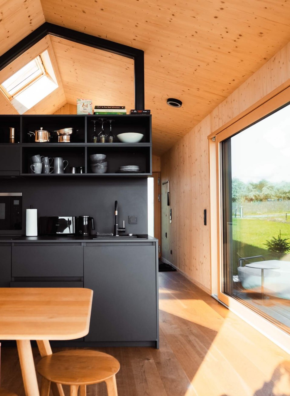 Modern eingerichtetes Tiny House mit schwarzer Küchenzeile, Holzmöbeln und einer großen Schiebetür zur Terrasse. // Stilvoller Minimalismus trifft auf Natur – Tiny House mit Panoramafenster, moderner Küche und gemütlichem Interior für eine entspannte Auszeit. // © mari.linni || Nicola Link Berlin Modern eingerichtetes Tiny House mit schwarzer Küchenzeile, Holzmöbeln und einer großen Schiebetür zur Terrasse.
