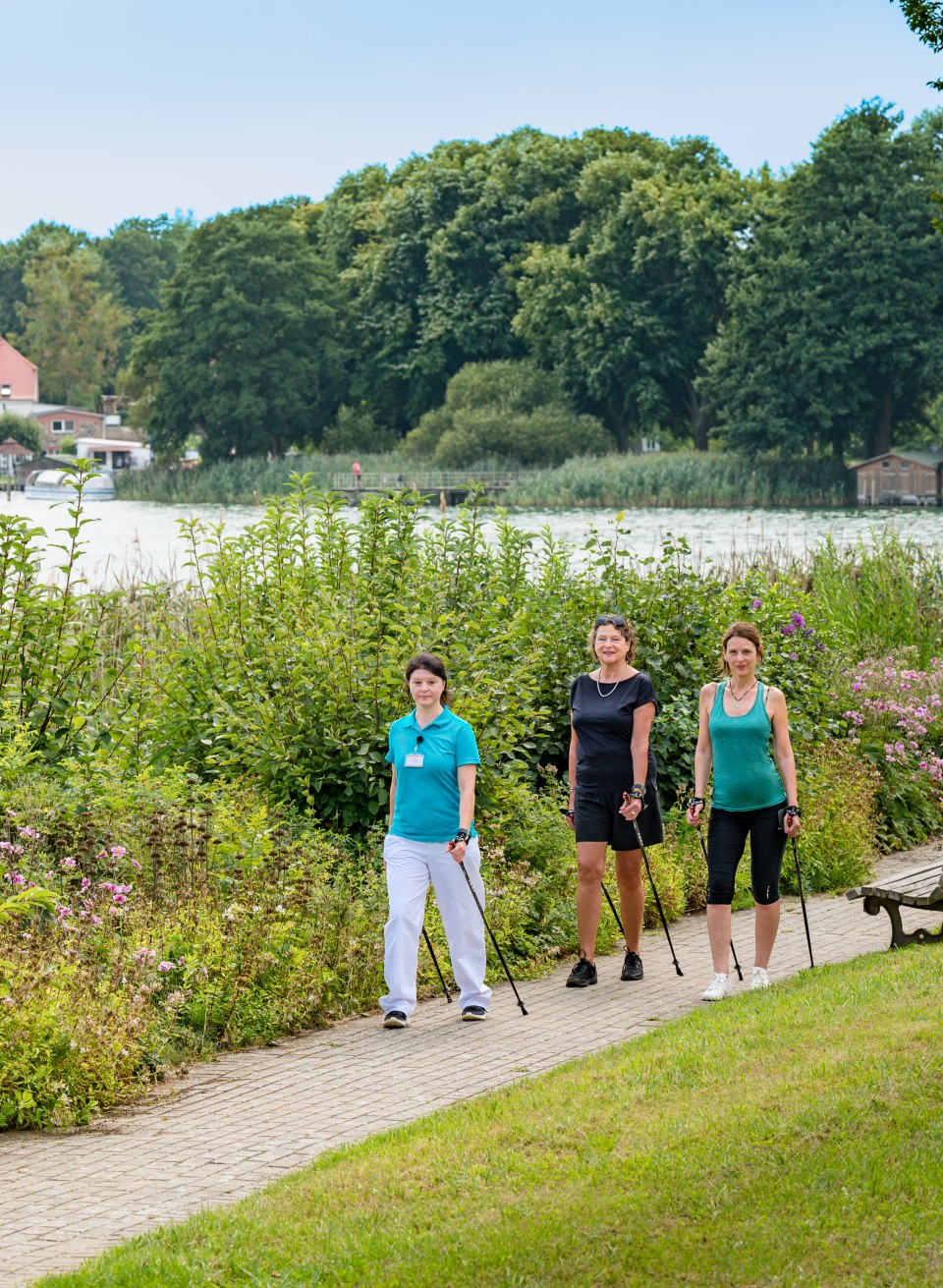 Die Nordic Walking-Kurse finden im Park direkt am See statt., © TMV/Tiemann Drei Frauen machen Nordic Walking im eigenen Park neben dem See des Medical Wellness Zentrums.