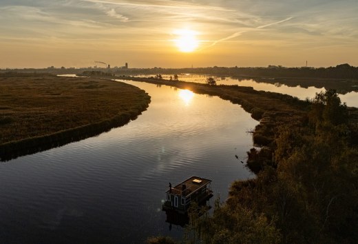 En husb&aring;d p&aring; Peene ved solnedgang, omgivet af et fredfyldt flodlandskab og gyldent lys i horisonten.