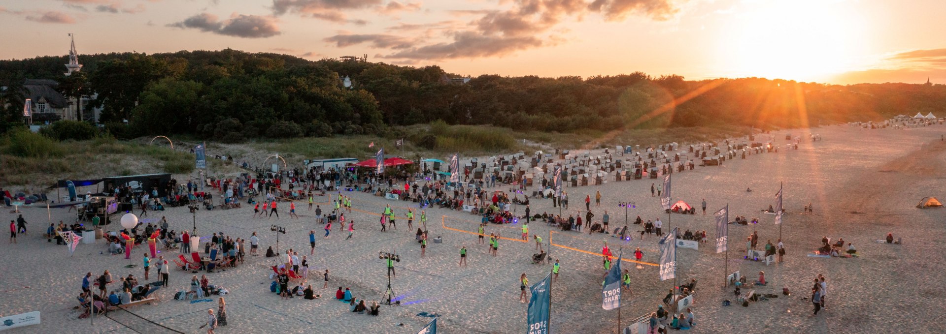 Mensen spelen glowvolleybal bij zonsondergang op het Oostzeestrand van de Kaiserb&auml;der tegen een achtergrond van duinen en strandstoelen.