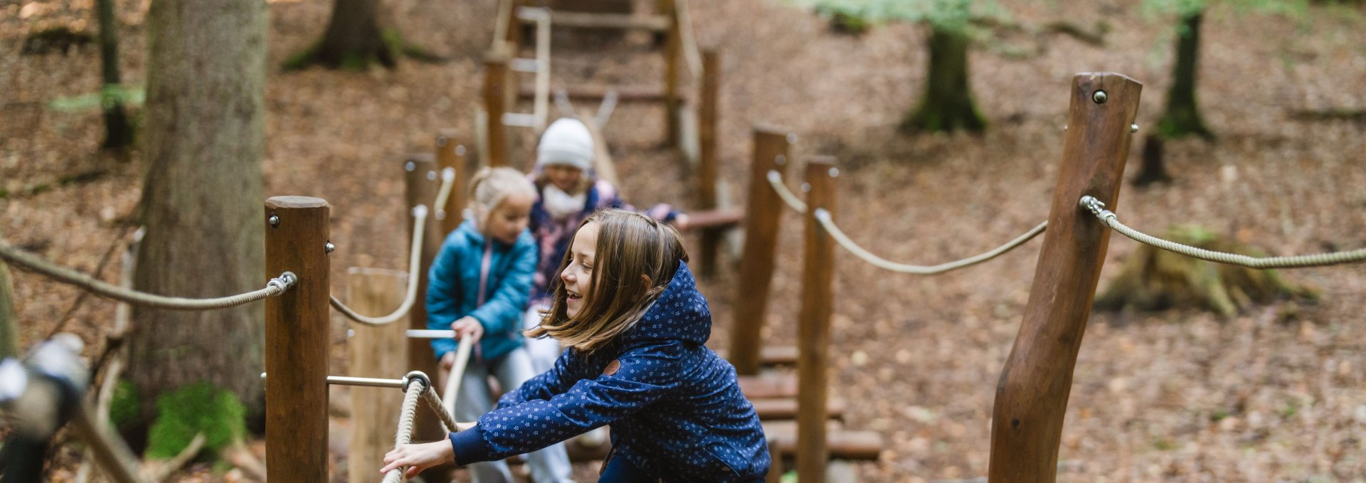 Een kind balanceert op een smalle balk in een speeltuin in het bos terwijl twee andere kinderen over het parcours op de achtergrond rennen.