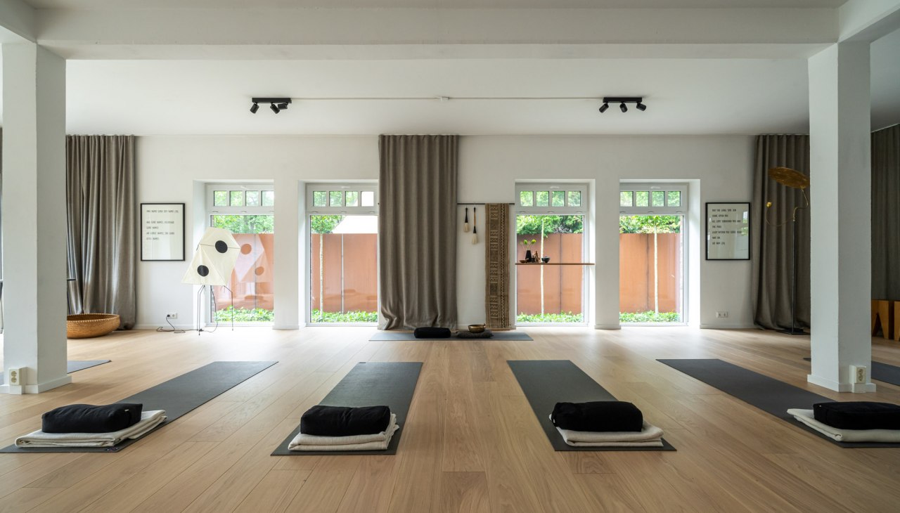 Ein moderner, minimalistischer Yoga- und Meditationsraum mit hellen Holzb&ouml;den, gro&szlig;en Fenstern und dezenter Dekoration. Die Matten sind bereits f&uuml;r eine bevorstehende Yoga-Sitzung vorbereitet, begleitet von weichen Kissen und Handt&uuml;chern., &copy; Hotel Carpe Diem