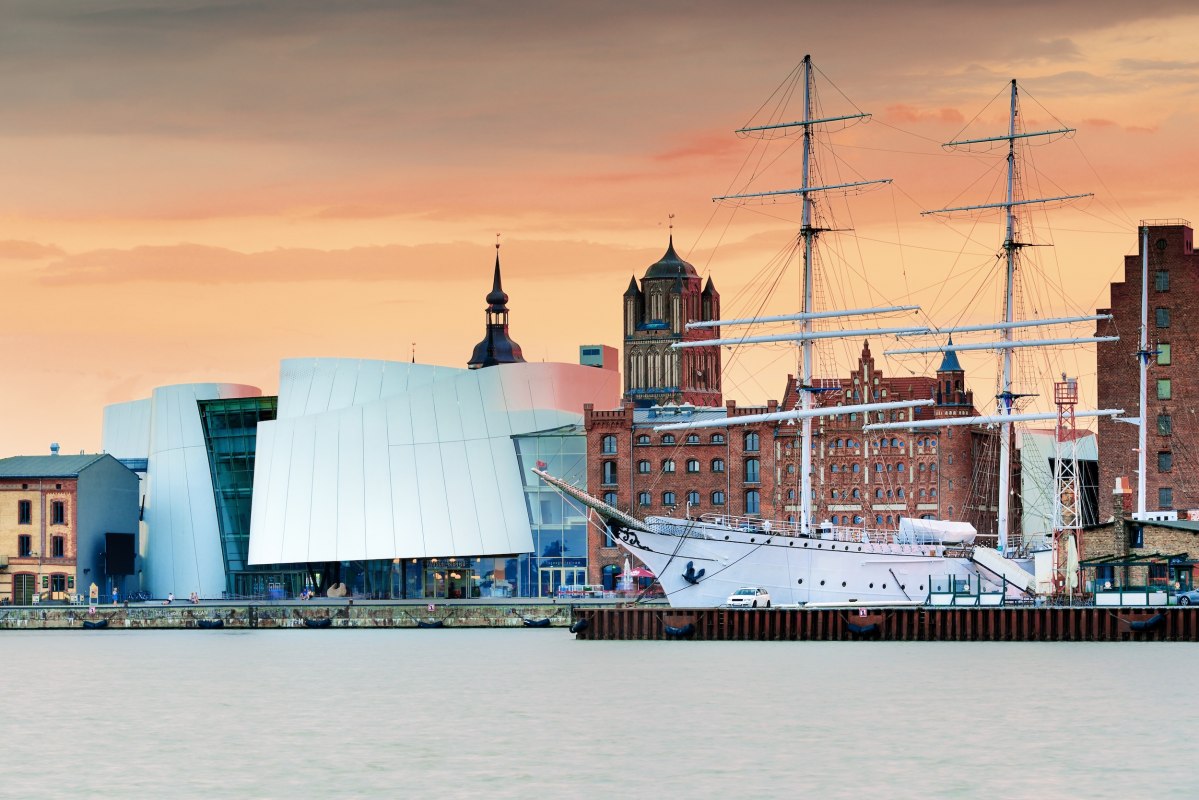Moderne Museumsarchitektur trifft auf hanseatisches Erbe: Das Ozeaneum am Stralsunder Hafen verschmilzt mit historischen Backsteinbauten und Segelschiffen zu einer einzigartigen Silhouette. Im Abendlicht leuchtet die UNESCO-Welterbestadt in warmen Farbtönen. Hier lässt sich maritime Geschichte hautnah erleben – vom Museumsdeck bis zur Hafenpromenade. // © Francesco Carovillano Moderne Museumsarchitektur trifft auf hanseatisches Erbe: Das Ozeaneum am Stralsunder Hafen verschmilzt mit historischen Backsteinbauten und Segelschiffen zu einer einzigartigen Silhouette. Im Abendlicht leuchtet die UNESCO-Welterbestadt in warmen Farbtönen. Hier lässt sich maritime Geschichte hautnah erleben – vom Museumsdeck bis zur Hafenpromenade. // © Francesco Carovillano