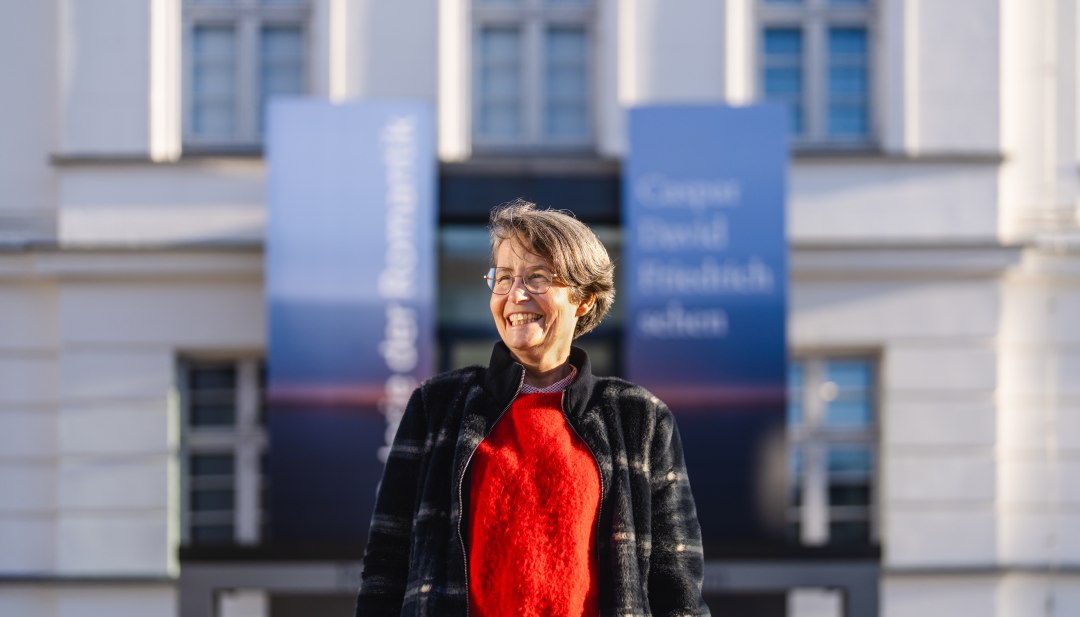 Een vrouw in een rode trui staat glimlachend voor de ingang van het Pommers Staatsmuseum in Greifswald.