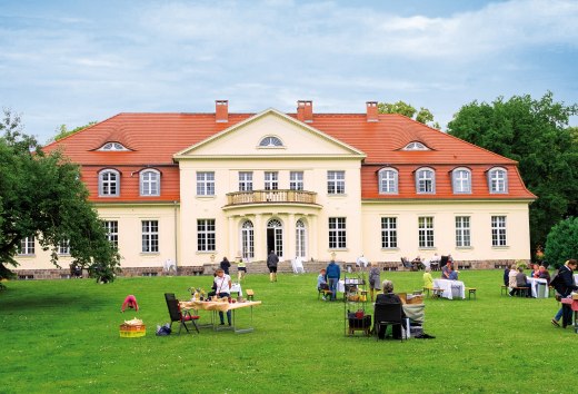 Menschen sitzen bei einer Mittsommerveranstaltung auf der Wiese vor dem Gutshaus Gevezin, w&auml;hrend das historische Geb&auml;ude hell im Tageslicht strahlt.