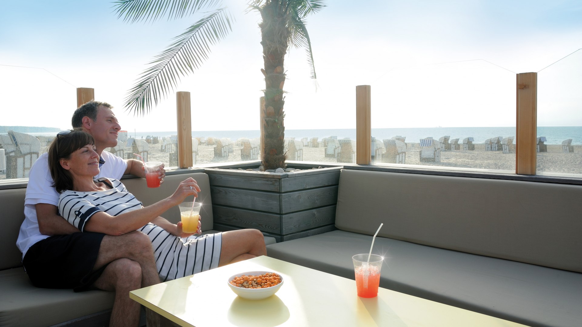 Paar entspannt mit Getr&auml;nken auf einer Lounge in der Strandbar D&Uuml;NE 13 in Warnem&uuml;nde, mit Blick auf Strand und Ostsee.