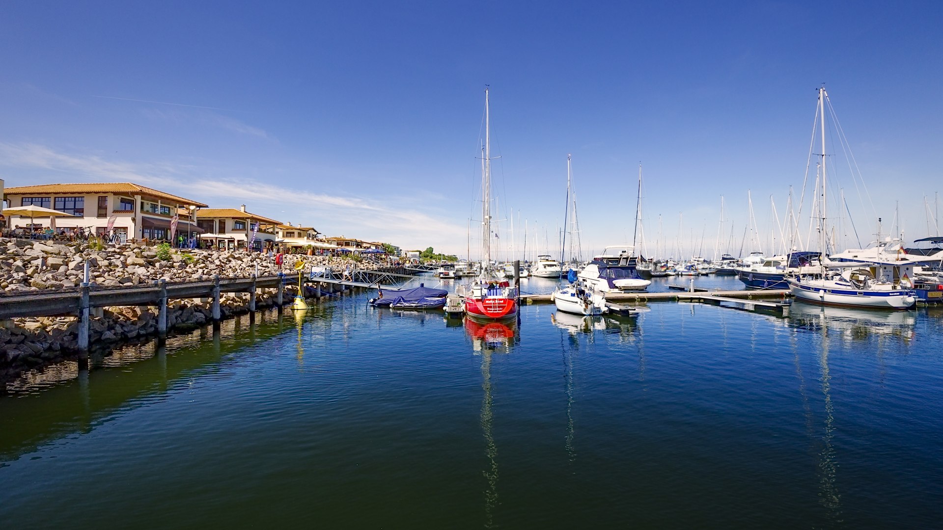 Blick auf den Yachthafen K&uuml;hlungsborn mit Segelbooten, Promenade und Restaurants bei klarem Himmel.