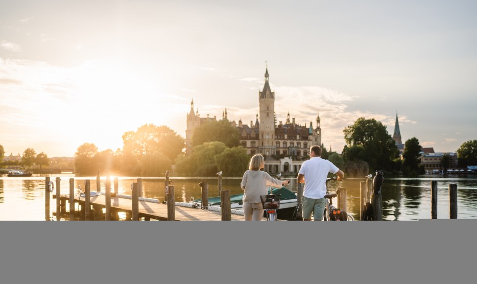 Ein Paar mit Fahrr&auml;dern steht auf einem Steg am Schweriner See und blickt im Abendlicht auf das Schweriner Schloss.