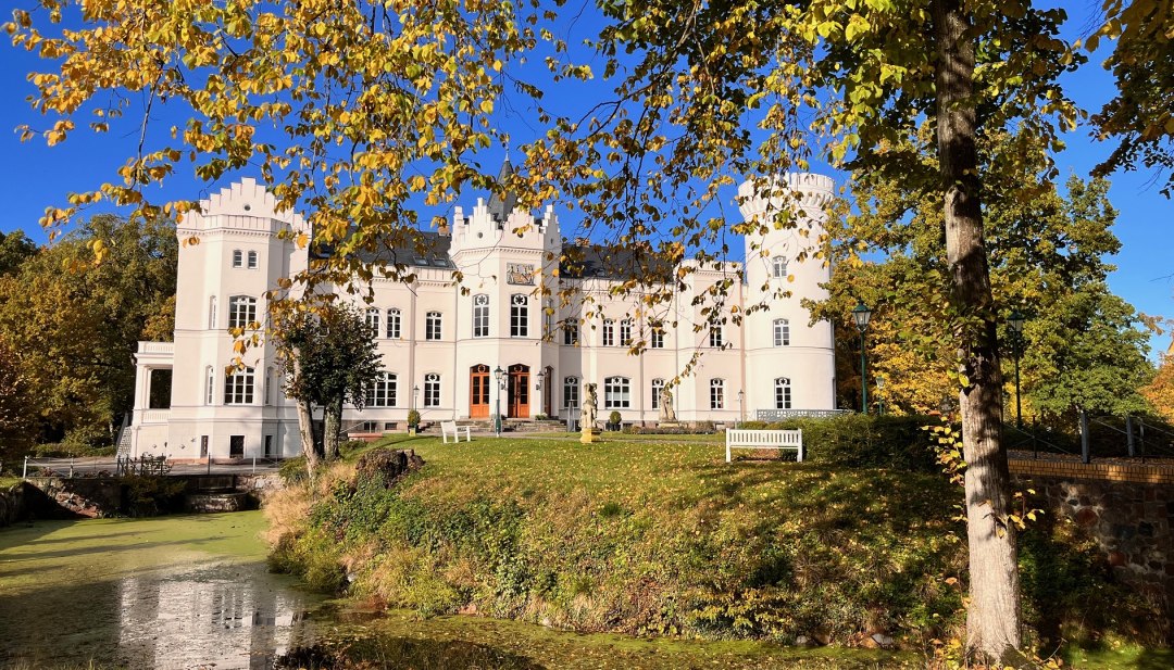 Voor kasteel Schlemmin schitteren herfstbladeren boven de gracht; de witte gevel steekt af tegen de blauwe lucht. // De witte gevel van het kasteel, het goudkleurige gebladerte, de stille slotgracht – Kasteel Schlemmin laat Mecklenburg-Vorpommern van zijn heldere kant zien. // © Schloss Schlemmin Voor kasteel Schlemmin schitteren herfstbladeren boven de gracht; de witte gevel steekt af tegen de blauwe lucht.