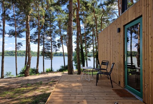 Entspannung pur im Wald – auf der Holzterrasse eines modernen Tiny Houses den Blick über den glitzernden See genießen und den Alltagsstress hinter sich lassen. Perfekt für einen naturnahen Urlaub inmitten grüner Wälder., © Ahoi Camp Canow Holzterrasse eines Tiny Houses mit Seeblick, umgeben von Kiefern in ruhiger Naturkulisse.