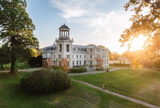 Schloss Kaarz in Mecklenburg-Schwerin im Sonnenuntergang mit Spaziergängern im gepflegten Schlosspark. // Abendstimmung in Mecklenburg-Schwerin – Schloss Kaarz erstrahlt im warmen Licht der untergehenden Sonne und lädt zu entspannten Spaziergängen durch die historische Parklandschaft ein. // © MV-T/Gross Schloss Kaarz in Mecklenburg-Schwerin im Sonnenuntergang mit Spaziergängern im gepflegten Schlosspark.