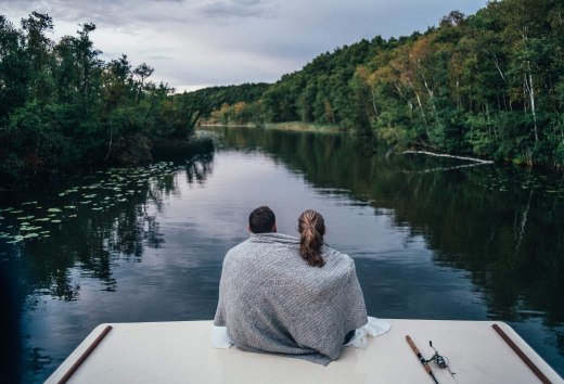 Ein Paar sitzt auf einem Hausboot, eingeh&uuml;llt in eine Decke, und blickt auf einen ruhigen, bewaldeten Fluss in der Mecklenburgischen Seenplatte.