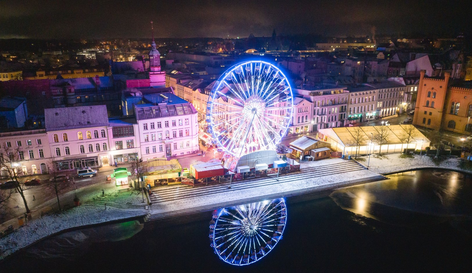 Beleuchtetes Riesenrad am Schweriner Pfaffenteich mit winterlichem Weihnachtsmarkt und Spiegelung im Wasser bei Nacht.