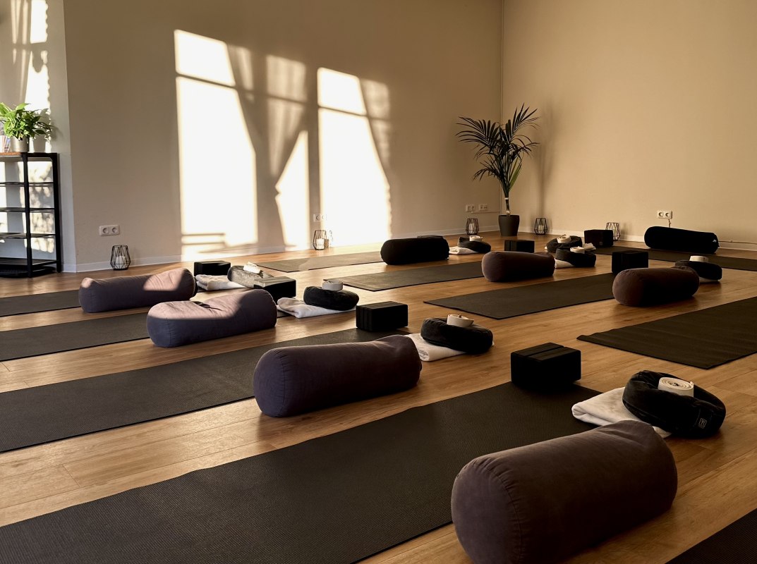 Lichte, minimalistische yogaruimte met houten vloer, verschillende yogamatten, meditatiekussens, bolsters en yogablokken. Warm zonlicht stroomt binnen door de grote ramen en cre&euml;ert een rustige, ontspannen sfeer voor yoga, meditatie en ontspanning. // &copy; MOVA PLACE - Yoga & Co. Studio