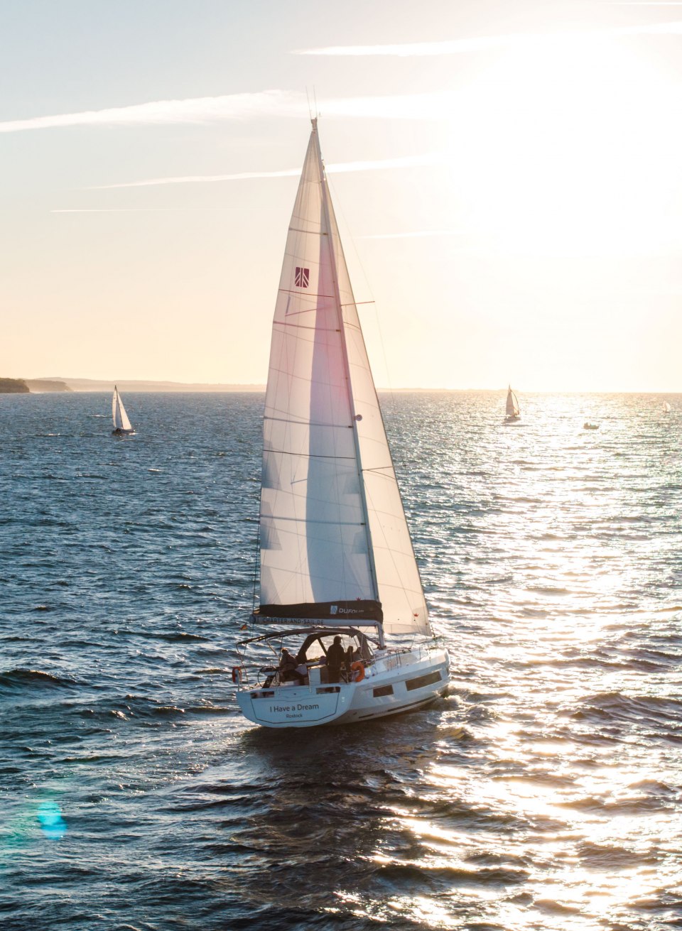 Segelboot mit wei&szlig;em Segel auf der Ostsee vor Warnem&uuml;nde bei Sonnenuntergang mit glitzerndem Wasser.