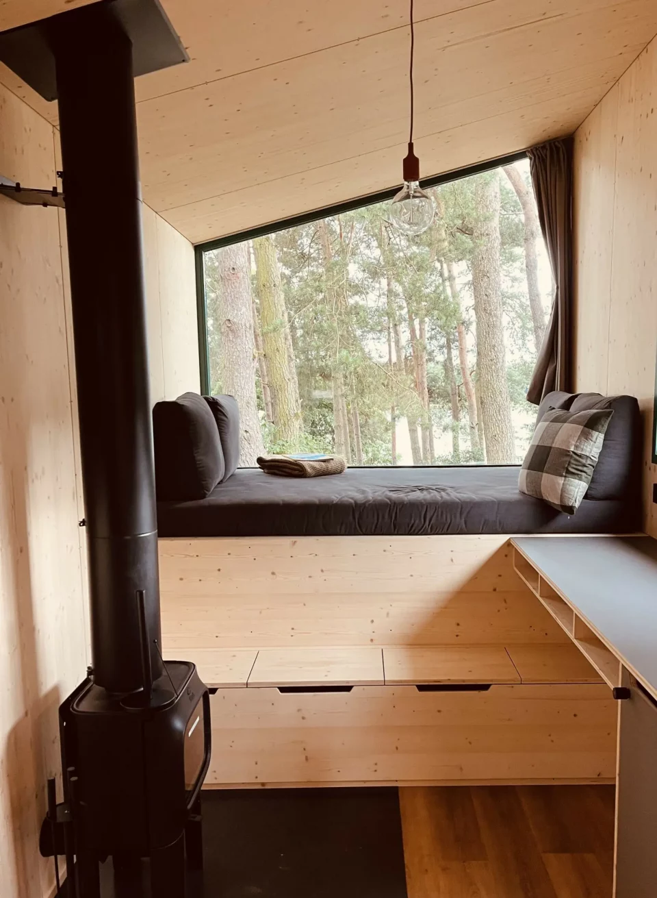Gemütlicher Innenraum eines Tiny Houses mit Holzverkleidung, Kaminofen und einem großen Panoramafenster mit Waldblick. // Minimalistisch wohnen, maximal entspannen – stilvolle Tiny Houses aus Holz bieten naturnahen Komfort für eine besondere Auszeit. // © Ahoi Camp Canow Gemütlicher Innenraum eines Tiny Houses mit Holzverkleidung, Kaminofen und einem großen Panoramafenster mit Waldblick.