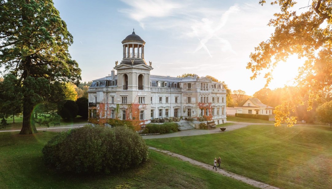 Schloss Kaarz in Mecklenburg-Schwerin im Sonnenuntergang mit Spaziergängern im gepflegten Schlosspark. // Abendstimmung in Mecklenburg-Schwerin – Schloss Kaarz erstrahlt im warmen Licht der untergehenden Sonne und lädt zu entspannten Spaziergängen durch die historische Parklandschaft ein. // © MV-T/Gross Schloss Kaarz in Mecklenburg-Schwerin im Sonnenuntergang mit Spaziergängern im gepflegten Schlosspark.
