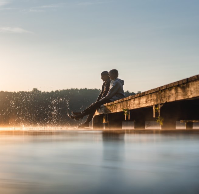 Zwei Personen sitzen im Sonnenaufgang auf einem Steg am Mirower See und lassen die F&uuml;&szlig;e ins glatte Wasser tauchen, w&auml;hrend feiner Nebel &uuml;ber der Oberfl&auml;che schwebt.