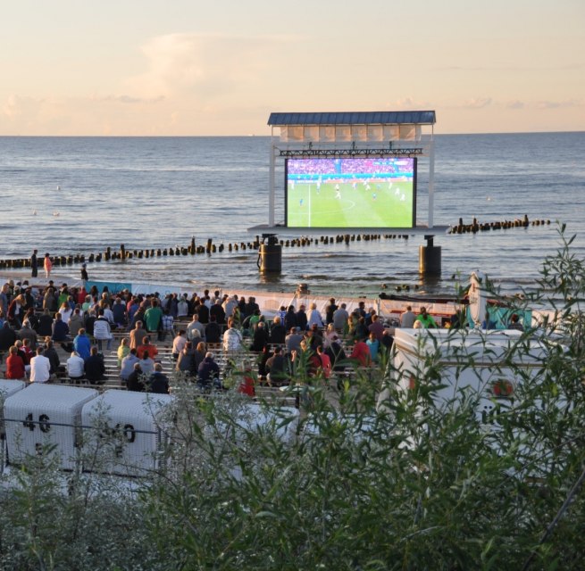 Eine große Menge schaut am Strand auf eine Leinwand im Wasser, dahinter liegt das ruhige Meer. // Fußball läuft auf der Großleinwand, das Publikum sitzt am Meer und teilt den Moment bis javascript://in den Abend. // © Eigenbetrieb Kaiserbäder Insel Usedom Eine große Menge schaut am Strand auf eine Leinwand im Wasser, dahinter liegt das ruhige Meer.