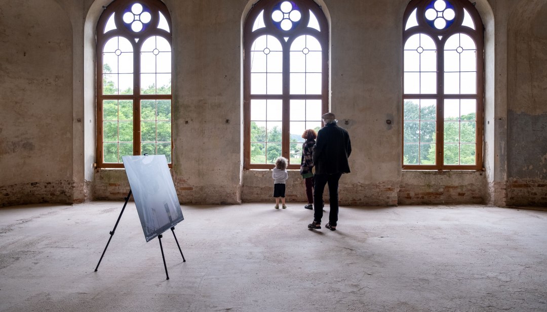 Familie betrachtet Ausstellung in historischem Saal mit gotischen Fenstern im Schloss Broock. // Licht fällt durch gotische Bogenfenster in die Räume von Schloss Broock. Zwischen freiliegendem Mauerwerk und klaren Linien eröffnen sich neue Perspektiven – Geschichte wird hier greifbar und lädt Generationen zum gemeinsamen Entdecken ein. // © Lars Schönherr Familie betrachtet Ausstellung in historischem Saal mit gotischen Fenstern im Schloss Broock.