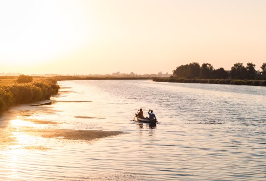 Zwei Personen paddeln in einem Kanu &uuml;ber einen ruhigen Fluss bei Sonnenuntergang, mit goldenem Licht auf dem Wasser.