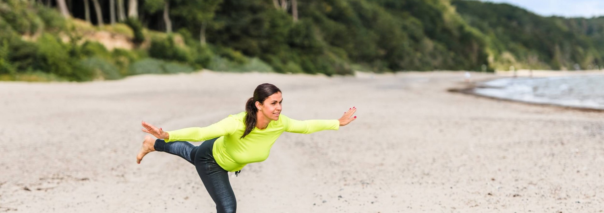 Eine Frau macht die "Standwaage" am Strand und hält das Gleichgewicht. // Stelle dich auf ein Bein, spanne die Rumpfmuskulatur an und ziehe die Schultern aktiv nach hinten unten. Beuge nun die Hüfte und senke den Oberkörper mit geradem Rücken nach vorne unten. Strecke dabei die Arme zur Seite oder nach vorne aus und achte darauf, die Spannung im Oberkörper nicht zu verlieren. Gleichzeitig führst du dein freies Bein gestreckt nach hinten bis Kopf, Oberkörper und Bein in einer horizontalen Linie liegen. Das Standbein kann leicht gebeugt sein. Halte die Position für einige Sekunden und richte dich dann wieder auf, mit Fokus auf die hintere Oberschenkelmuskulatur. Wichtig: der Rücken muss gerade bleiben; Halswirbel- und Wirbelsäule sollen eine Linie bilden. 6-10 Wiederholungen pro Seite // © MV-T/Tiemann Eine Frau macht die "Standwaage" am Strand und hält das Gleichgewicht.