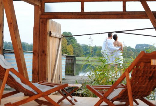 Ein Paar in Handtüchern steht auf einer Terrasse am See, umgeben von Schilf, mit Blick auf das Wasser in der Feldberger Seenlandschaft. Im Vordergrund sind Holzliegen zu sehen. // Entspannung pur: Ein Paar genießt den Blick auf den See nach einem Saunagang in der Feldberger Seenlandschaft. // © MV-T/Duerst Ein Paar in Handtüchern steht auf einer Terrasse am See, umgeben von Schilf, mit Blick auf das Wasser in der Feldberger Seenlandschaft. Im Vordergrund sind Holzliegen zu sehen.