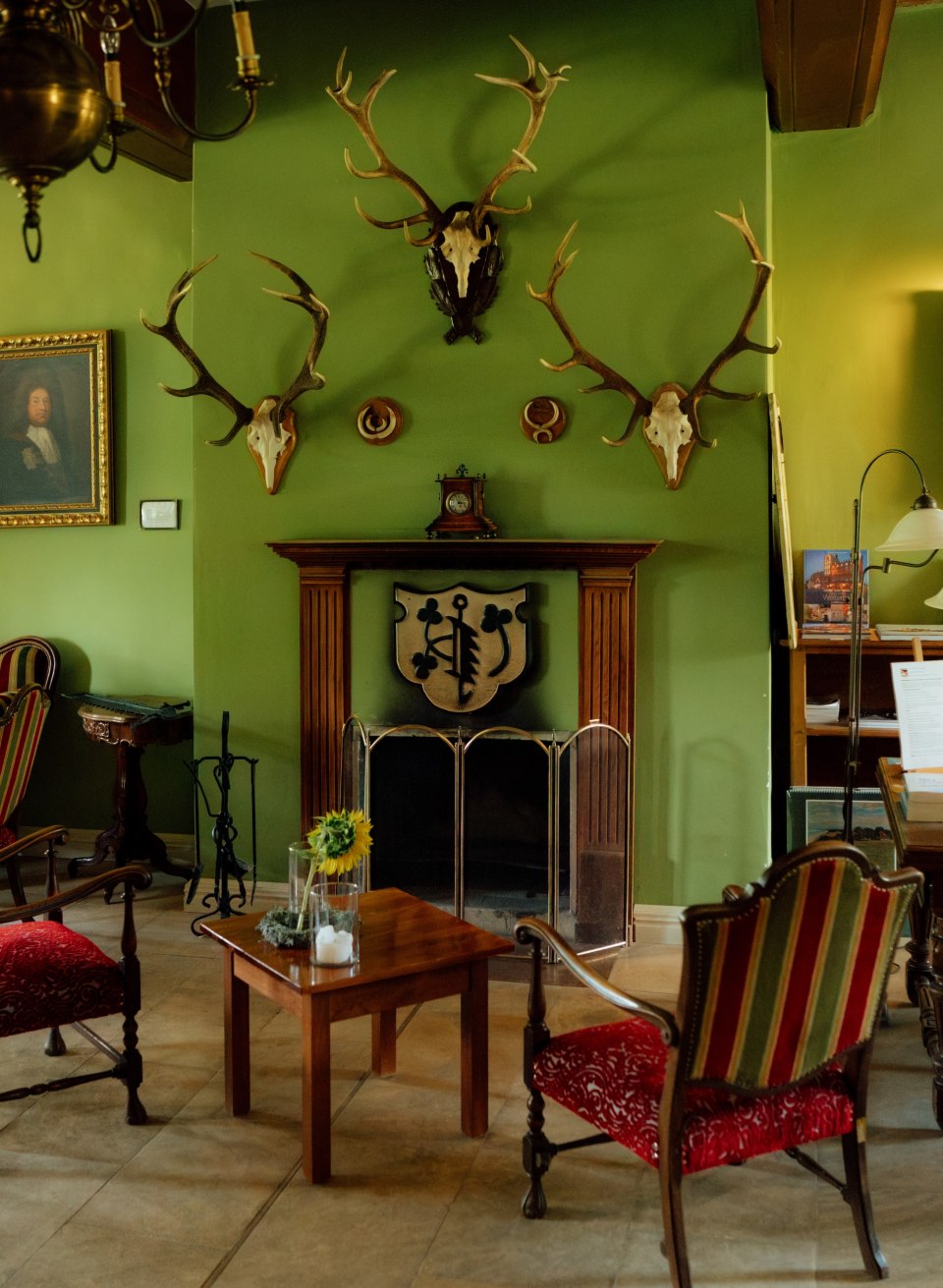 Kamin mit Holzrahmen an grüner Wand, darüber Geweihe und Wappen; davor stehen Sessel und ein Tisch im Gutshaus Ludorf. // Grün gestrichene Wände, alte Geweihe und ein Kamin als Mittelpunkt: Im Gutshaus Ludorf wartet ein Raum zum Runterkommen – drinnen ankommen in Mecklenburg-Vorpommern. // © MV-T/Petermann Kamin mit Holzrahmen an grüner Wand, darüber Geweihe und Wappen; davor stehen Sessel und ein Tisch im Gutshaus Ludorf.
