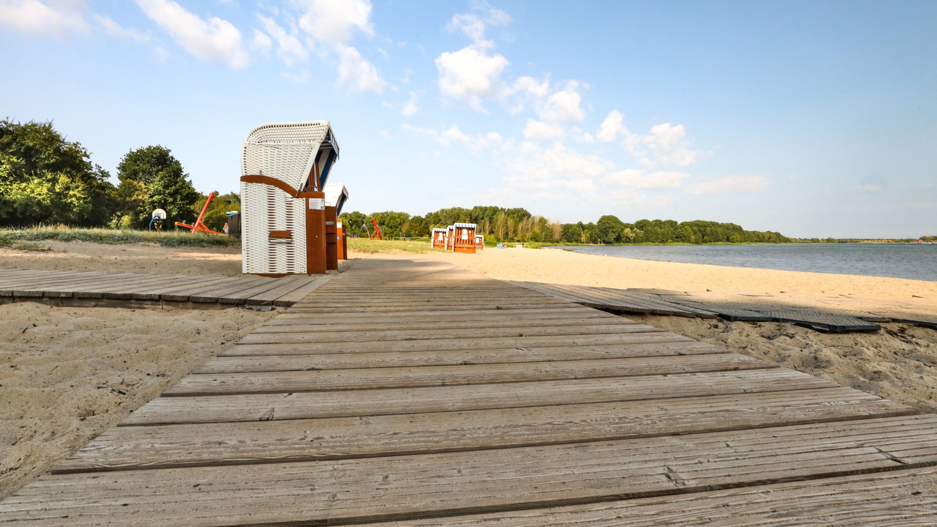 Holzsteg f&uuml;hrt barrierefrei zu Strandk&ouml;rben am Sandstrand des Schaalsees, umgeben von ruhiger Natur und blauem Himmel.