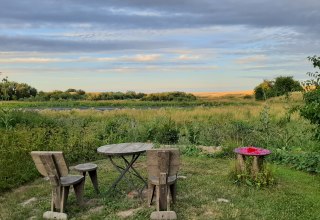 Der Sitzplatz am Weiher mit 2 Stühlen, 2 Hockern und 1 Tisch aus Holz. Der Blick geht über den Gartenrand und den zeitweise ausgetrockneten Weiher in die Landschaft. // © Mirabell e.V. Der Sitzplatz am Weiher mit 2 Stühlen, 2 Hockern und 1 Tisch aus Holz. Der Blick geht über den Gartenrand und den zeitweise ausgetrockneten Weiher in die Landschaft. // © Mirabell e.V.