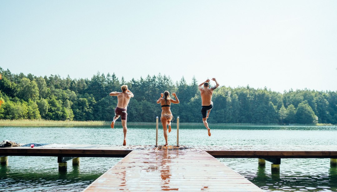 Drei junge Menschen springen gemeinsam von einem Holzsteg in einen klaren See, umgeben von dichtem Wald.