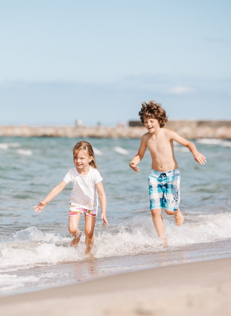 Zwei Kinder rennen lachend durch flache Ostseewellen am Strand, im Hintergrund steht ein gr&uuml;ner Leuchtturm auf der Mole.