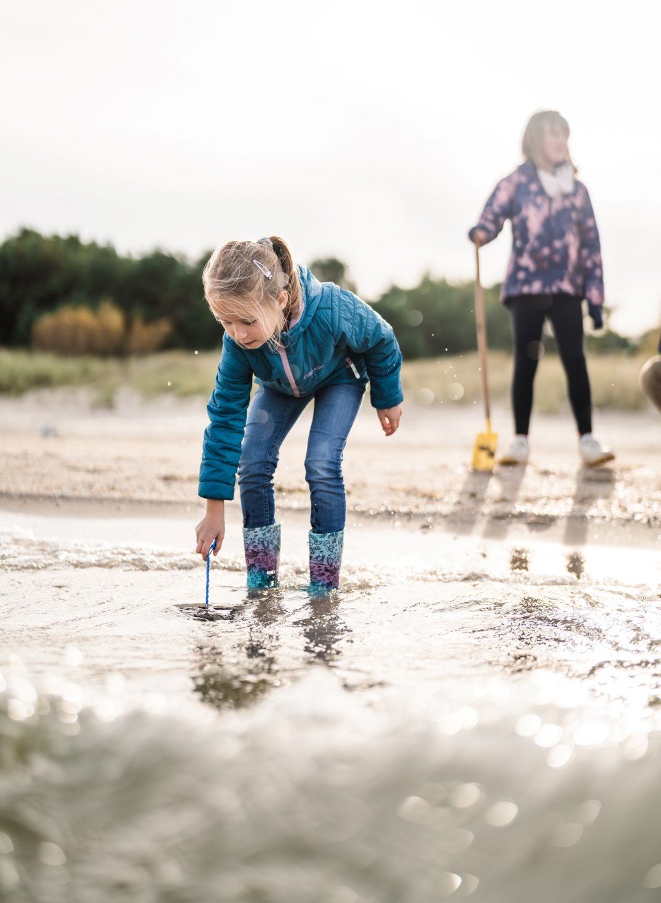 Ein M&auml;dchen in Gummistiefeln steht geb&uuml;ckt im Wasser am Strand und sucht nach etwas im Sand, w&auml;hrend ein weiteres Kind im Hintergrund steht.