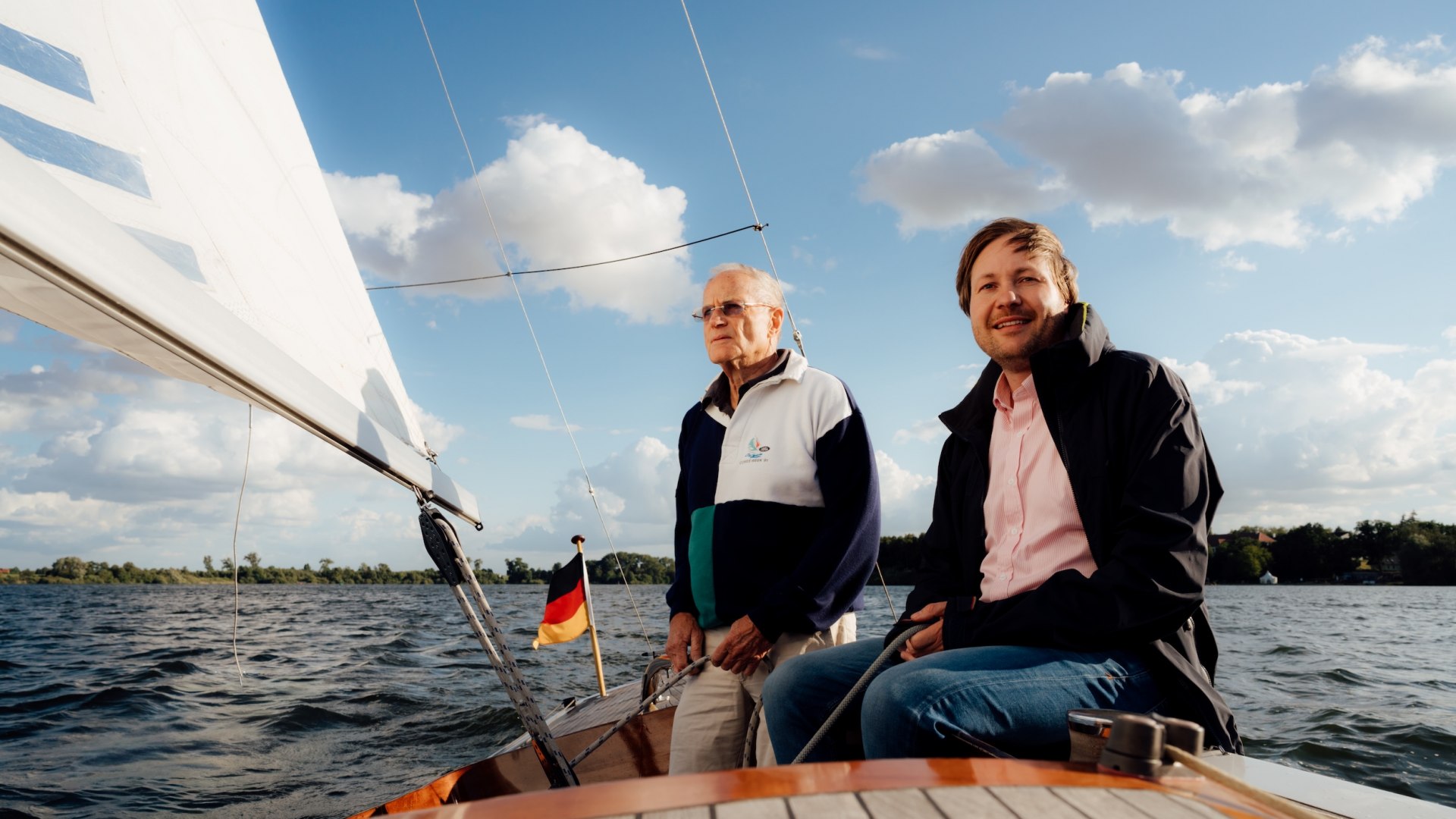 Jannis und Manfred Achtenhagen sitzen auf einem Segelboot auf der Müritz, Segel und Wolkenhimmel im Hintergrund. // Wind im Segel, Weite vor dem Bug: Jannis Achtenhagen sitzt mit Manfred Achtenhagen an Bord und lässt die Müritz arbeiten – klarer Kurs // © MV-T/Petermann Jannis und Manfred Achtenhagen sitzen auf einem Segelboot auf der Müritz, Segel und Wolkenhimmel im Hintergrund.