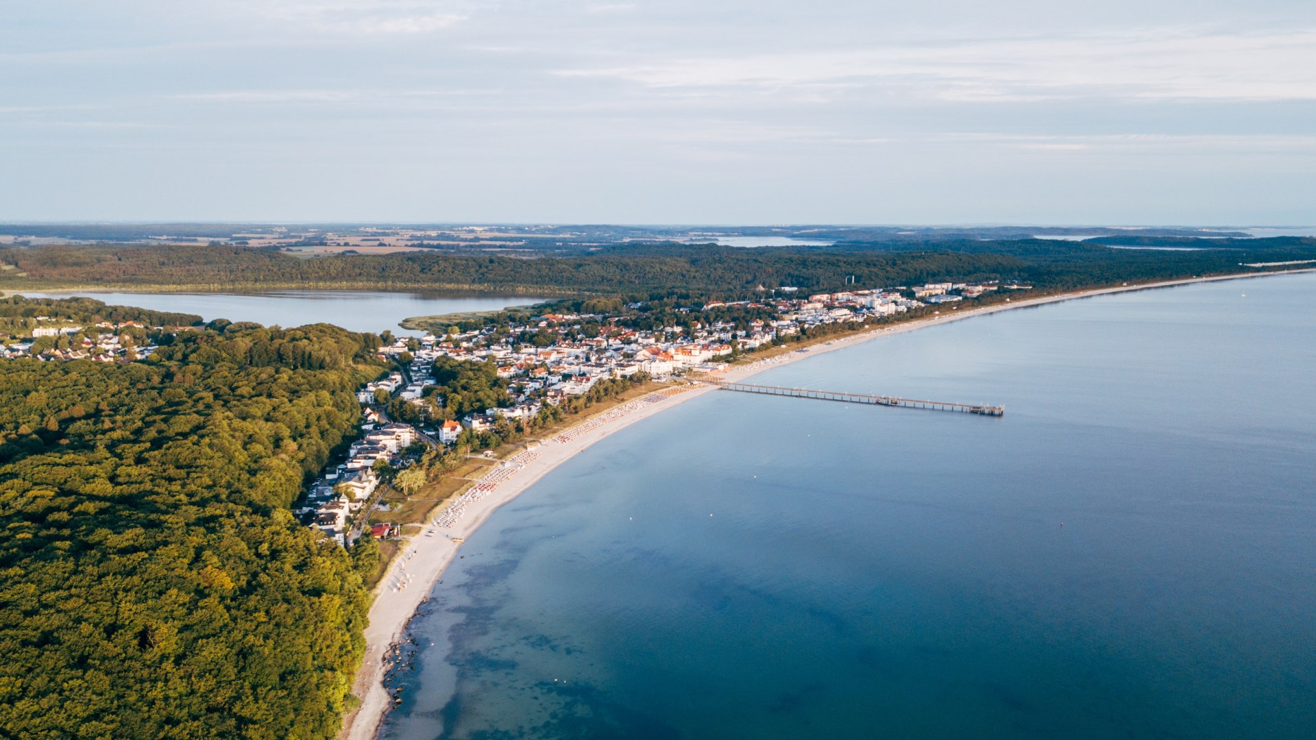 Luftaufnahme des Ostseebads Binz auf Rügen, das sich entlang der geschwungenen Küstenlinie erstreckt. Die Seebrücke ragt weit ins ruhige, türkisfarbene Wasser, während der Ort von dichten Wäldern und Seen umgeben ist. Der Himmel ist klar und die friedliche Landschaft lädt zum Verweilen ein., © TMV/Friedrich Luftaufnahme von Binz auf Rügen mit Seebrücke, Küstenlinie und umliegenden Wäldern.