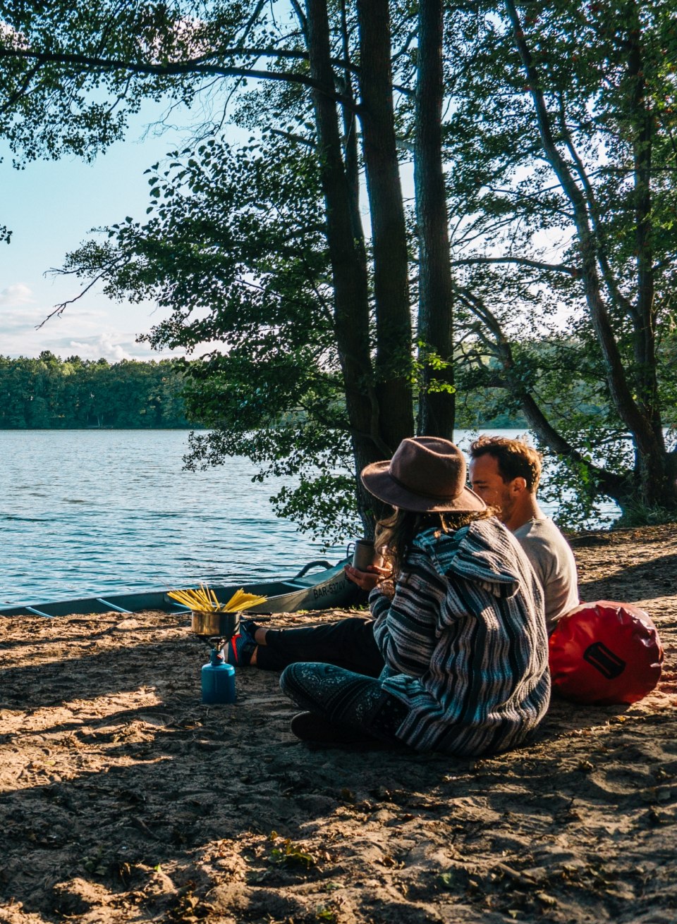 Ein Paar sitzt am Wasser mit einem Gaskocher und kocht Nudeln am Campingplatz Hexenw&auml;ldchen