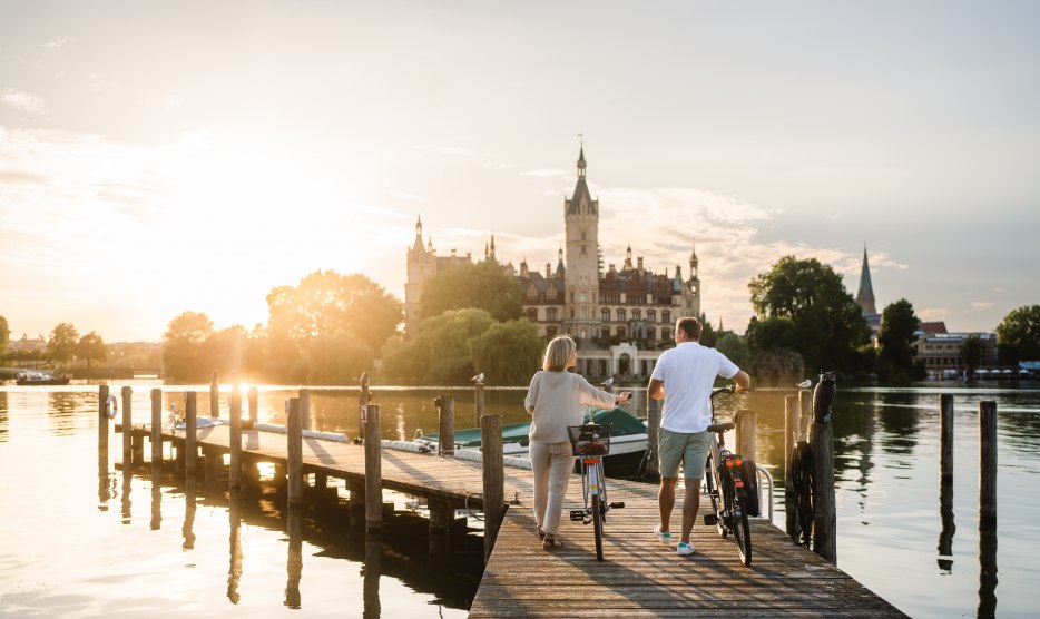 Ein Paar mit Fahrr&auml;dern steht auf einem Steg am Schweriner See und blickt im Abendlicht auf das Schweriner Schloss.