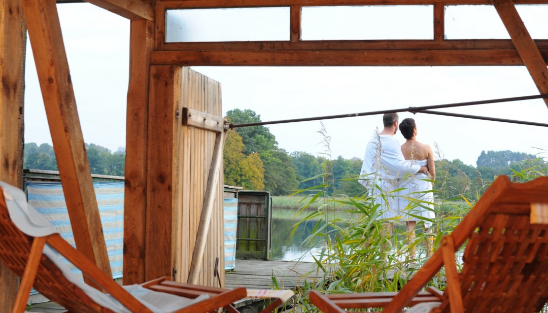 Ein Paar in Handt&uuml;chern steht auf einer Terrasse am See, umgeben von Schilf, mit Blick auf das Wasser in der Feldberger Seenlandschaft. Im Vordergrund sind Holzliegen zu sehen.