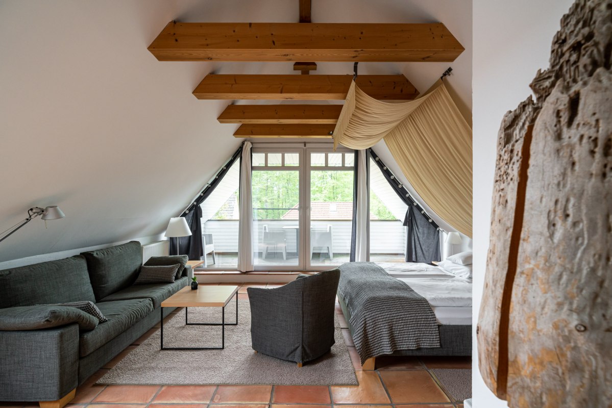 Ein stilvoll eingerichtetes Dachzimmer mit sichtbaren Holzbalken und einem gem&uuml;tlichen Ambiente. Der Raum bietet eine komfortable Sitzecke, ein gro&szlig;es Bett und Zugang zu einem Balkon mit Blick ins Gr&uuml;ne. Warme Terrakotta-B&ouml;den und helle Stoffe runden das gem&uuml;tliche Design ab und laden zum Entspannen ein. // &copy; Hotel Carpe Diem