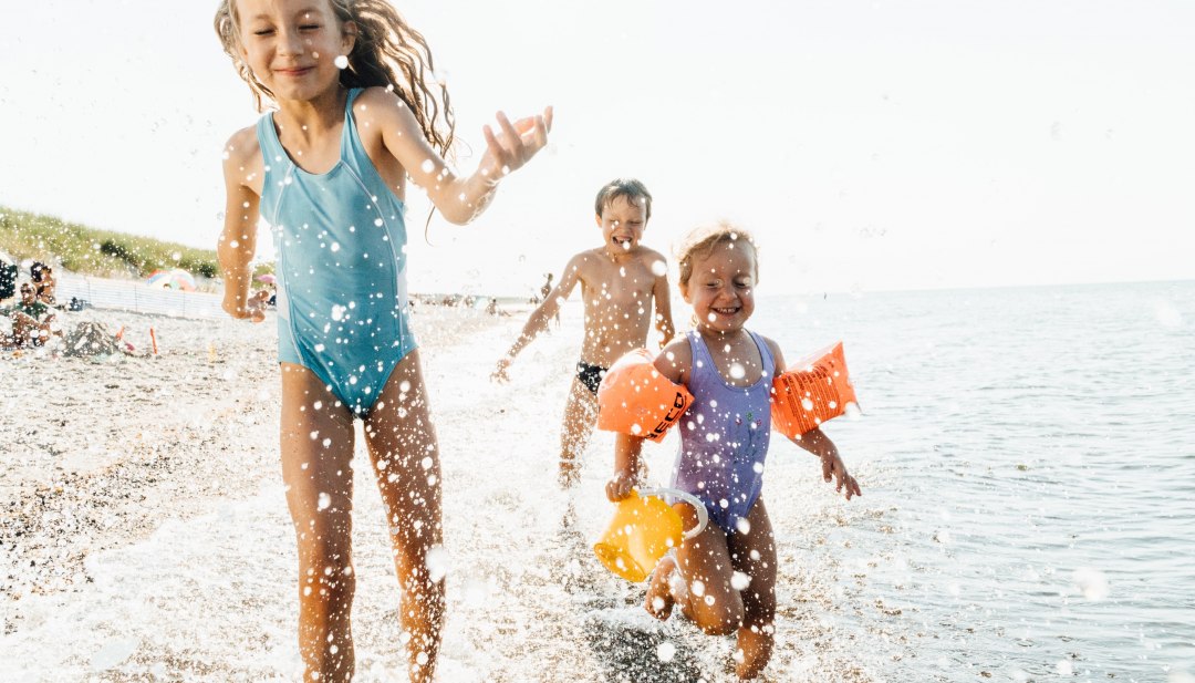 Drei Kinder laufen lachend durch seichtes Ostseewasser am Strand von Dierhagen, Spritzwasser im Vordergrund.