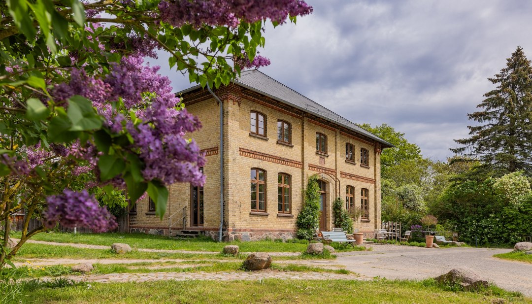 Gutshaus Lexow mit blühendem Flieder im Vordergrund und historischer Backsteinfassade. // Violette Fliederblüten rahmen das Gutshaus Lexow ein, während sich der Himmel über der historischen Fassade auftürmt. Zwischen Grün und Blütenpracht atmet der Ort Ruhe – ein Platz zum Ankommen und Durchatmen. // © Gutshaus Lexow Gutshaus Lexow mit blühendem Flieder im Vordergrund und historischer Backsteinfassade.