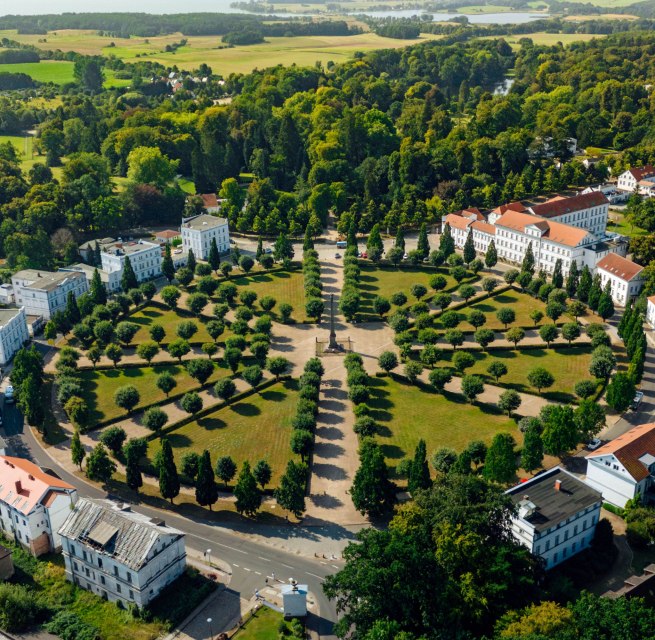 Luftaufnahme des Circus in Putbus auf R&uuml;gen, einem kreisf&ouml;rmigen Platz mit symmetrisch angeordneten B&auml;umen und klassizistischen Geb&auml;uden, umgeben von gr&uuml;ner Natur und l&auml;ndlicher Landschaft.